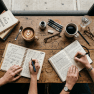 Two hands writing in separate notebooks on a wooden table with coffee cups, pens, and books