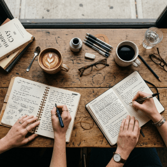 Two hands writing in separate notebooks on a wooden table with coffee cups, pens, and books